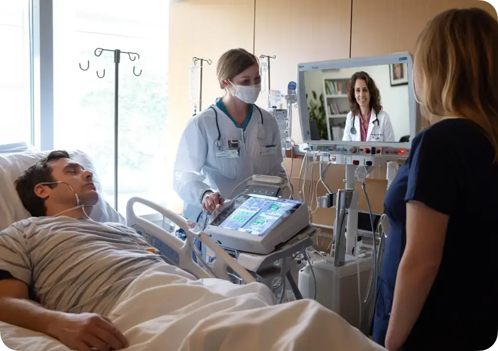 A team of nurses in an ICU talking to a teleneurologist on a tv screen, patient laying in bed